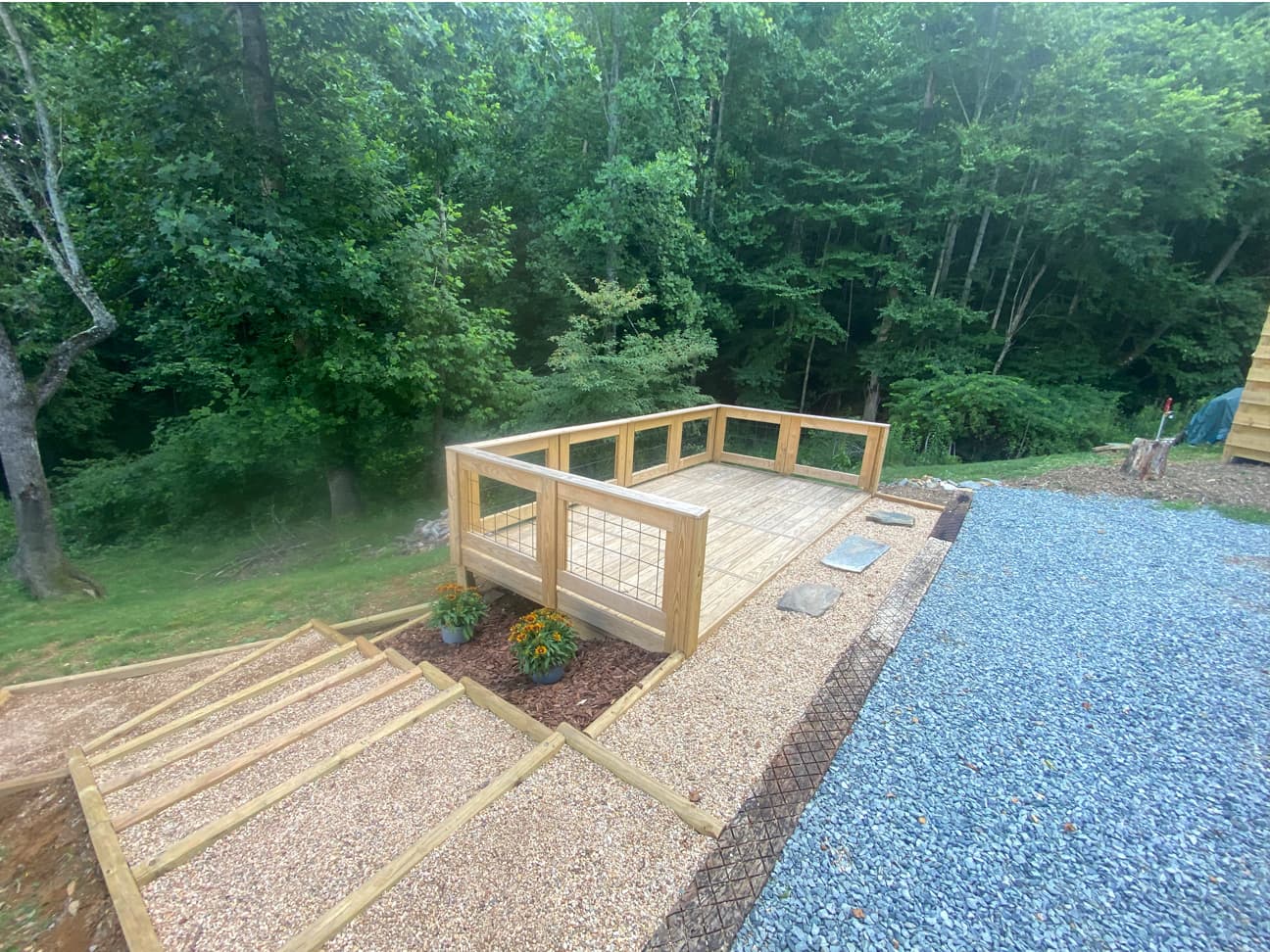 Close-up of a finished guest deck with wire-panel railing, wood mulch beds with potted flowers, and gravel steps leading up to the deck and driveway