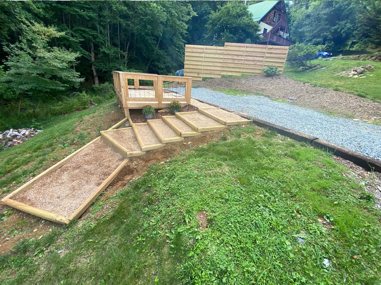 Side view of the completed wooden deck with tiered gravel steps leading down the hillside, a privacy fence panel, and a neighboring cabin with a green metal roof visible in the background