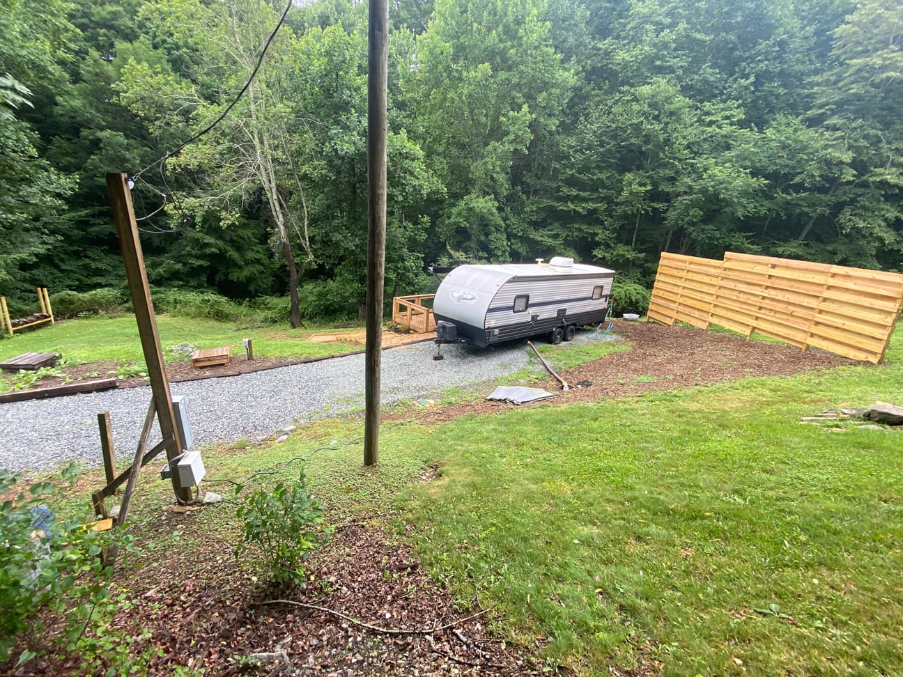 View of a travel trailer parked on the gravel pad next to the deck and privacy fence panel, with a wooded backgrop and green lawn in the background